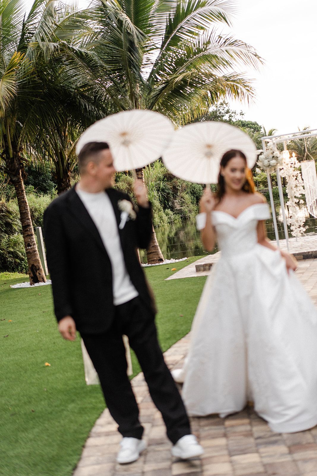 Bride and groom holding parasols walk on a path, smiles. The groom wears a black suit, white shirt, and sneakers. The bride wears a white dress.