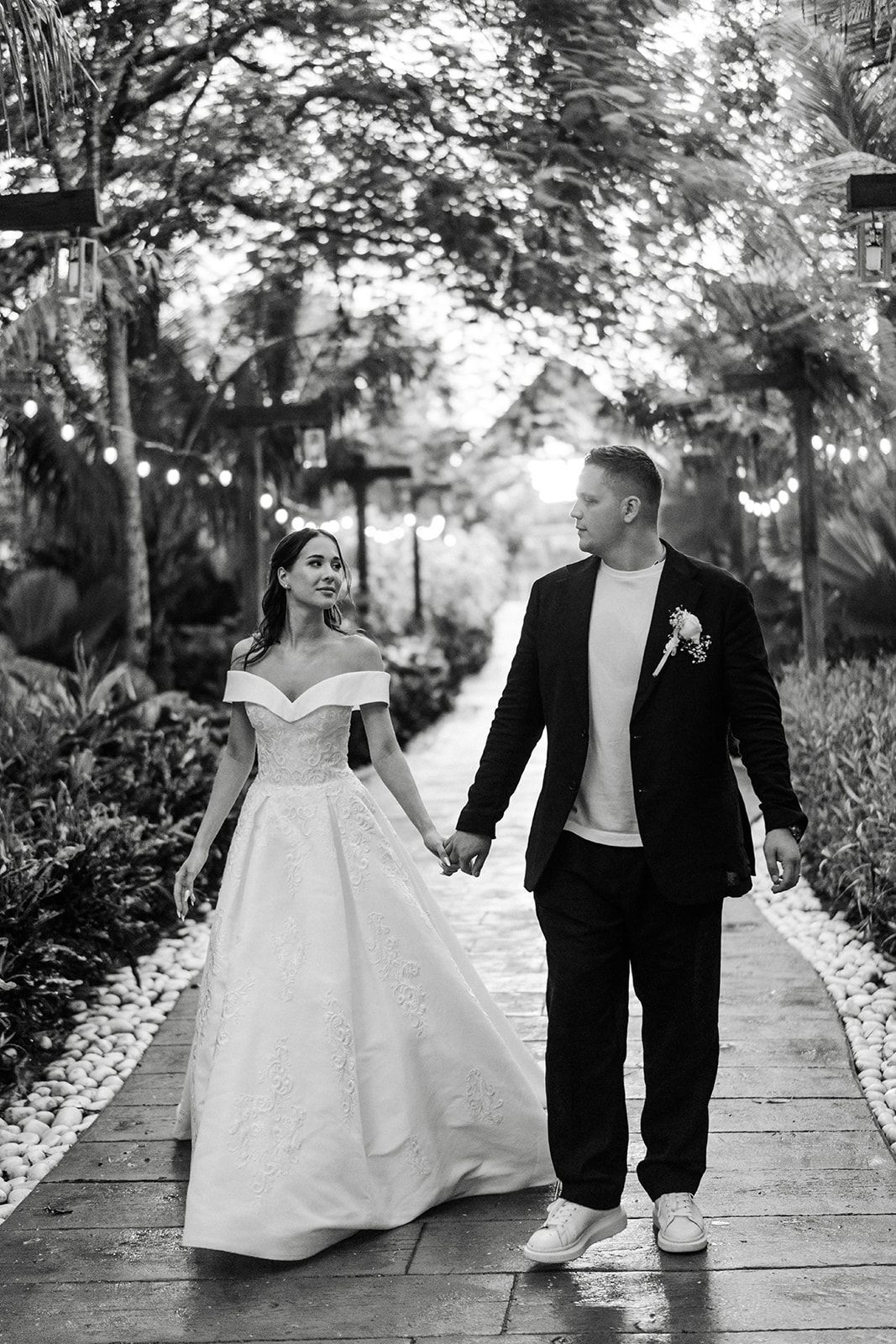 Wedding couple holding hands, walking along a stone path, looking at each other. Fairy lights in the background.