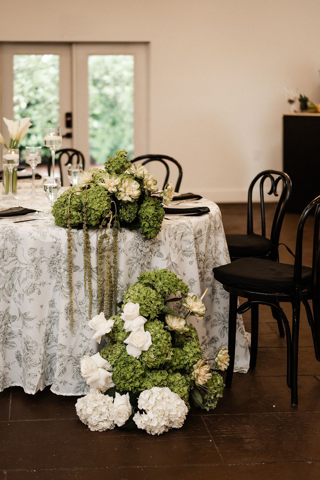 Round table set for a wedding, adorned with green and white floral arrangements. Black chairs and a patterned tablecloth.