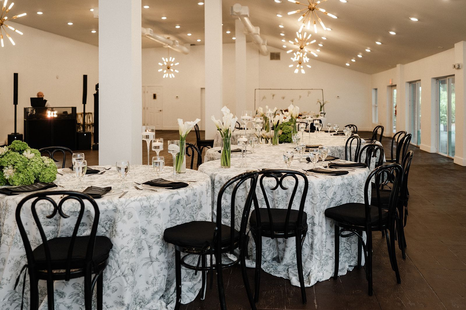 Elegant event space with tables set for a gathering, white floral tablecloths, black chairs, and modern lighting.