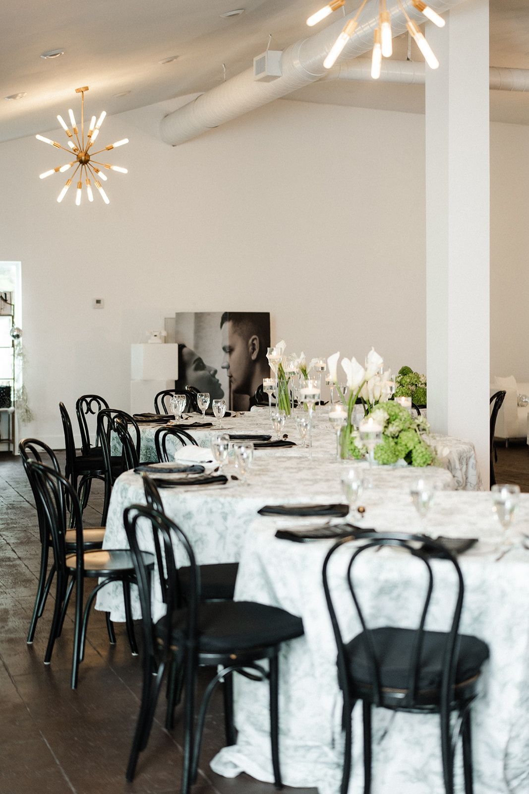 Elegant dining room with tables set for a meal; black chairs, white tablecloths, floral centerpieces.