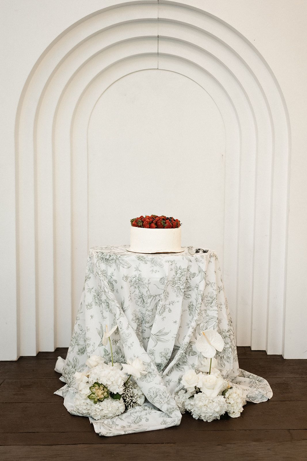 Cake on a table draped with patterned fabric, set before a white arched wall with flowers.