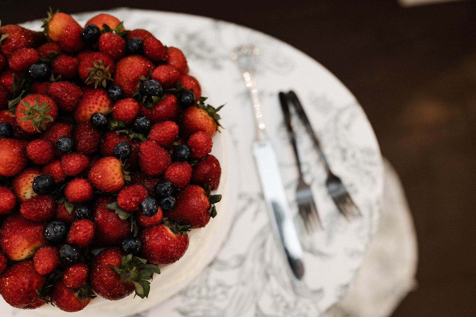 Cake topped with strawberries and blueberries on a table with a knife and forks.