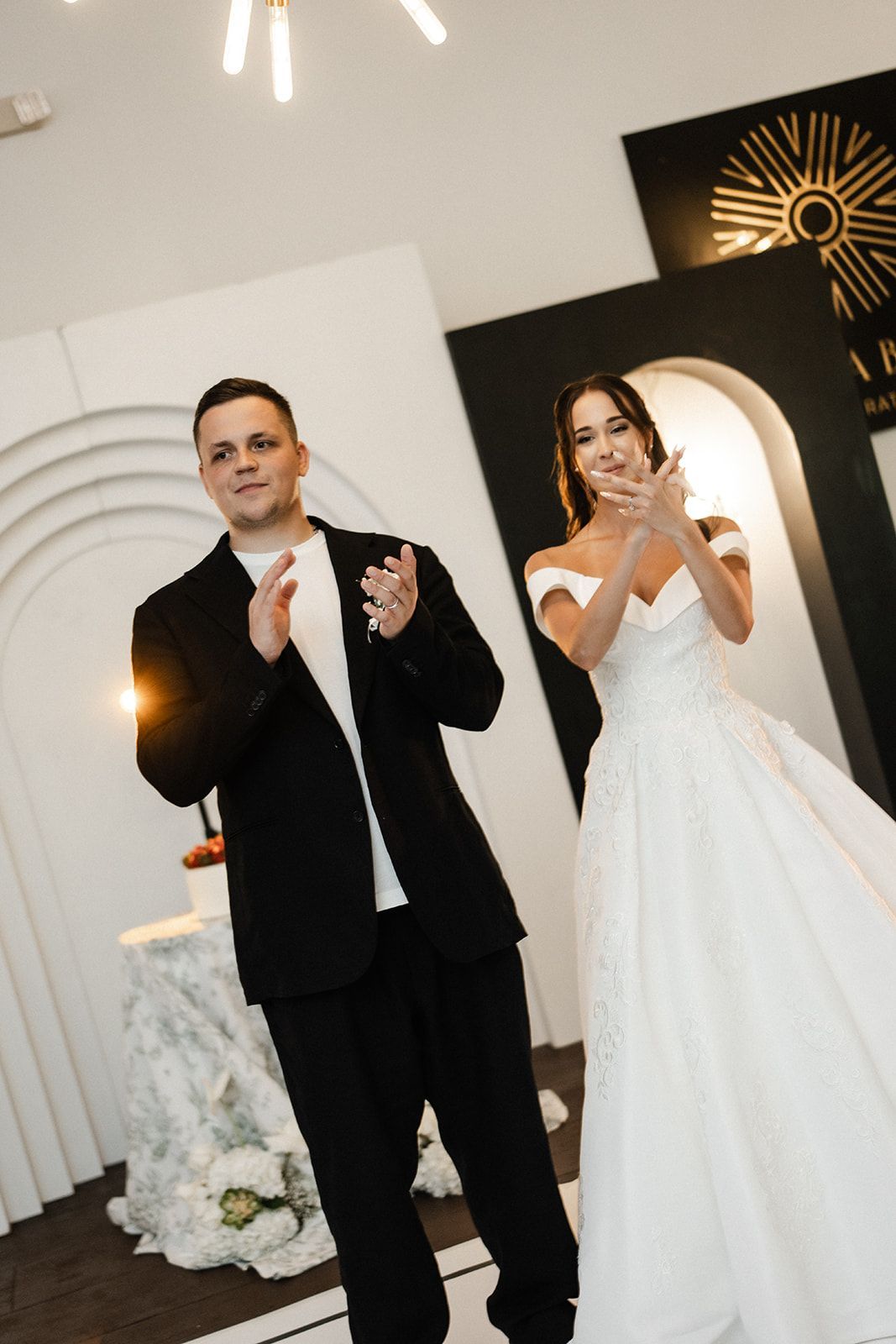 Bride and groom clapping at a reception. The woman wears a white gown, and the man a black suit.