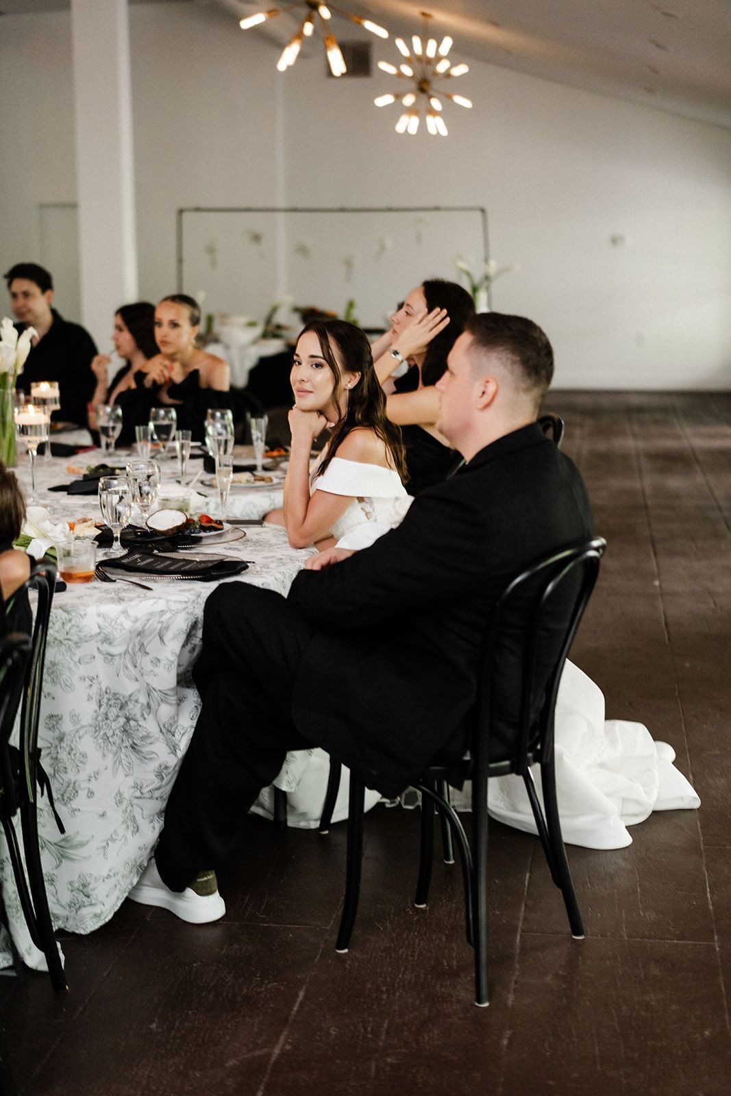Guests at a wedding reception seated at a decorated table, looking towards the speaker.