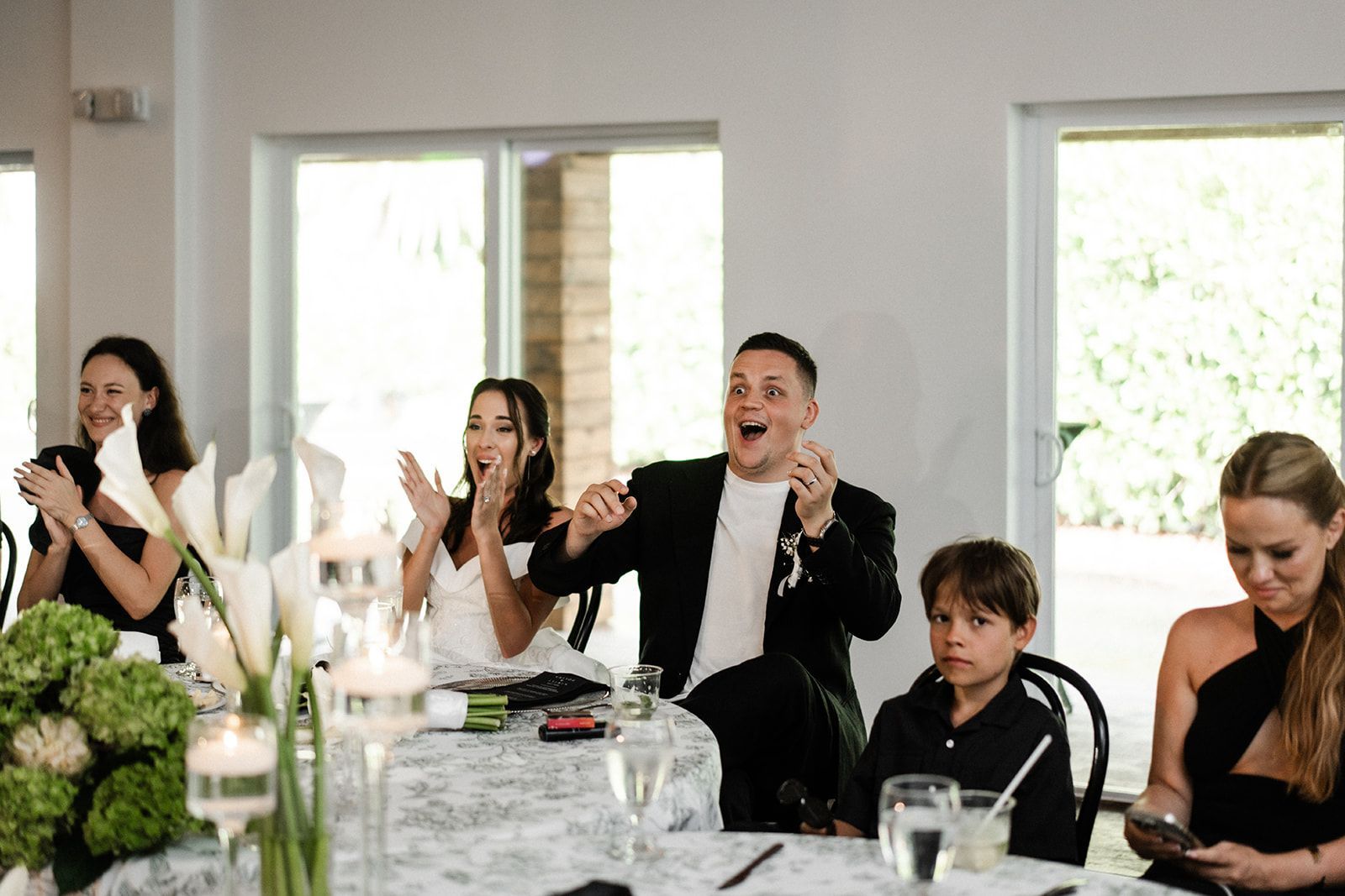 People clapping at a table, floral centerpieces. Man with excited expression.