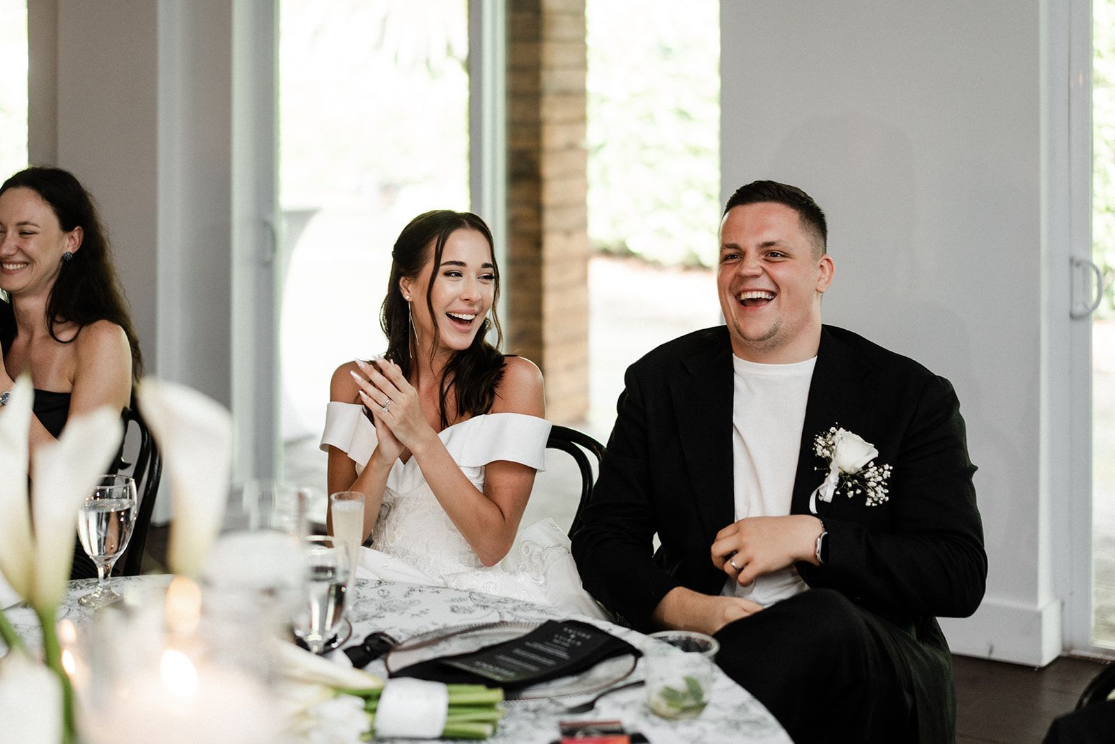 Bride and groom laughing at a wedding reception table. Bride applauds, groom chuckles.