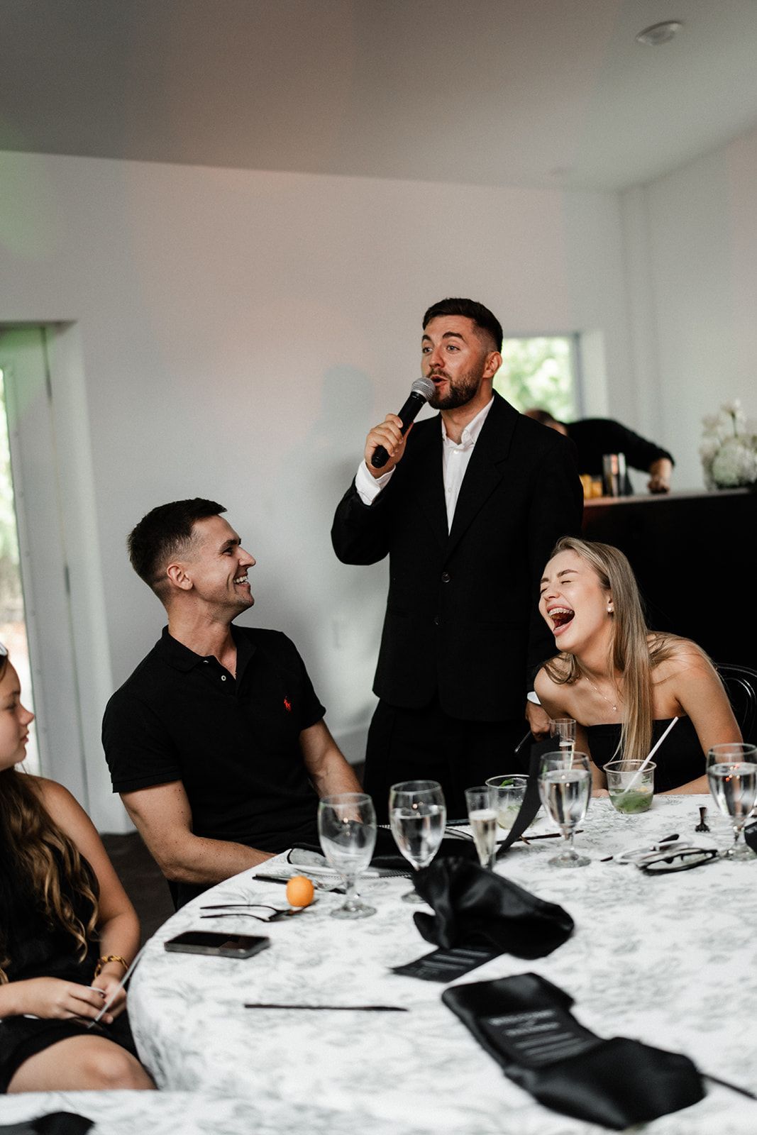 Man in suit speaks into microphone at a table. Guests laugh, drink and watch in a bright room.
