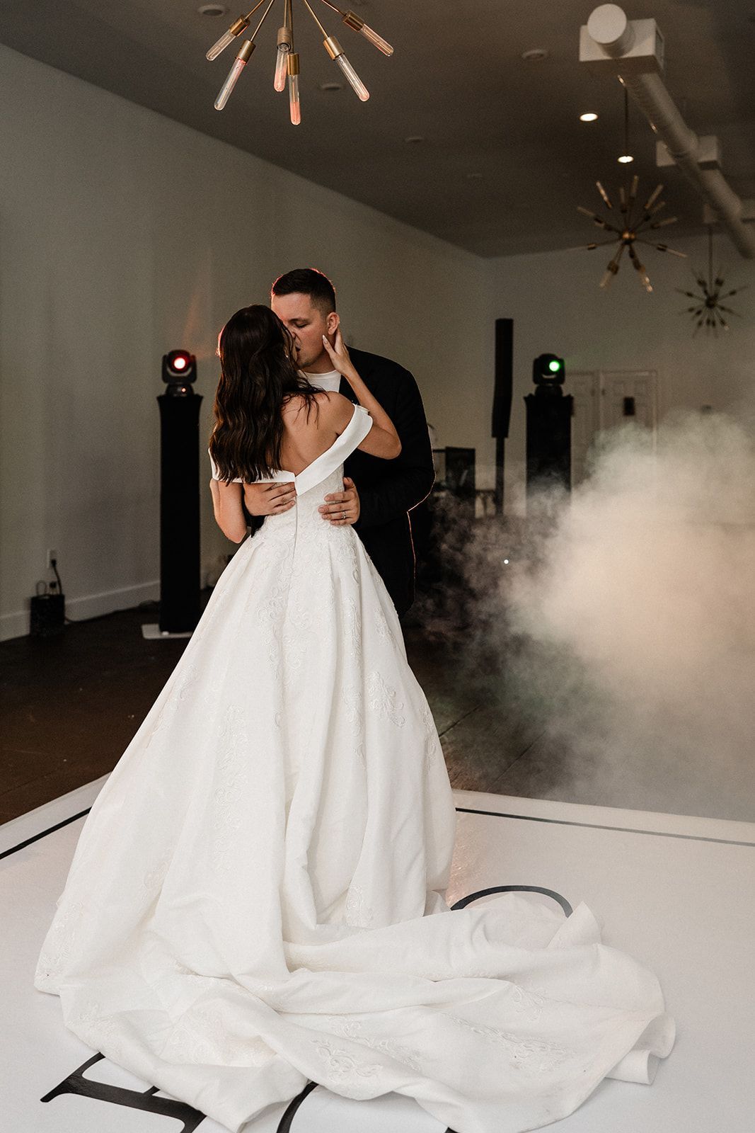Couple dancing at a wedding; bride in white gown, groom in black, on a dance floor with fog.