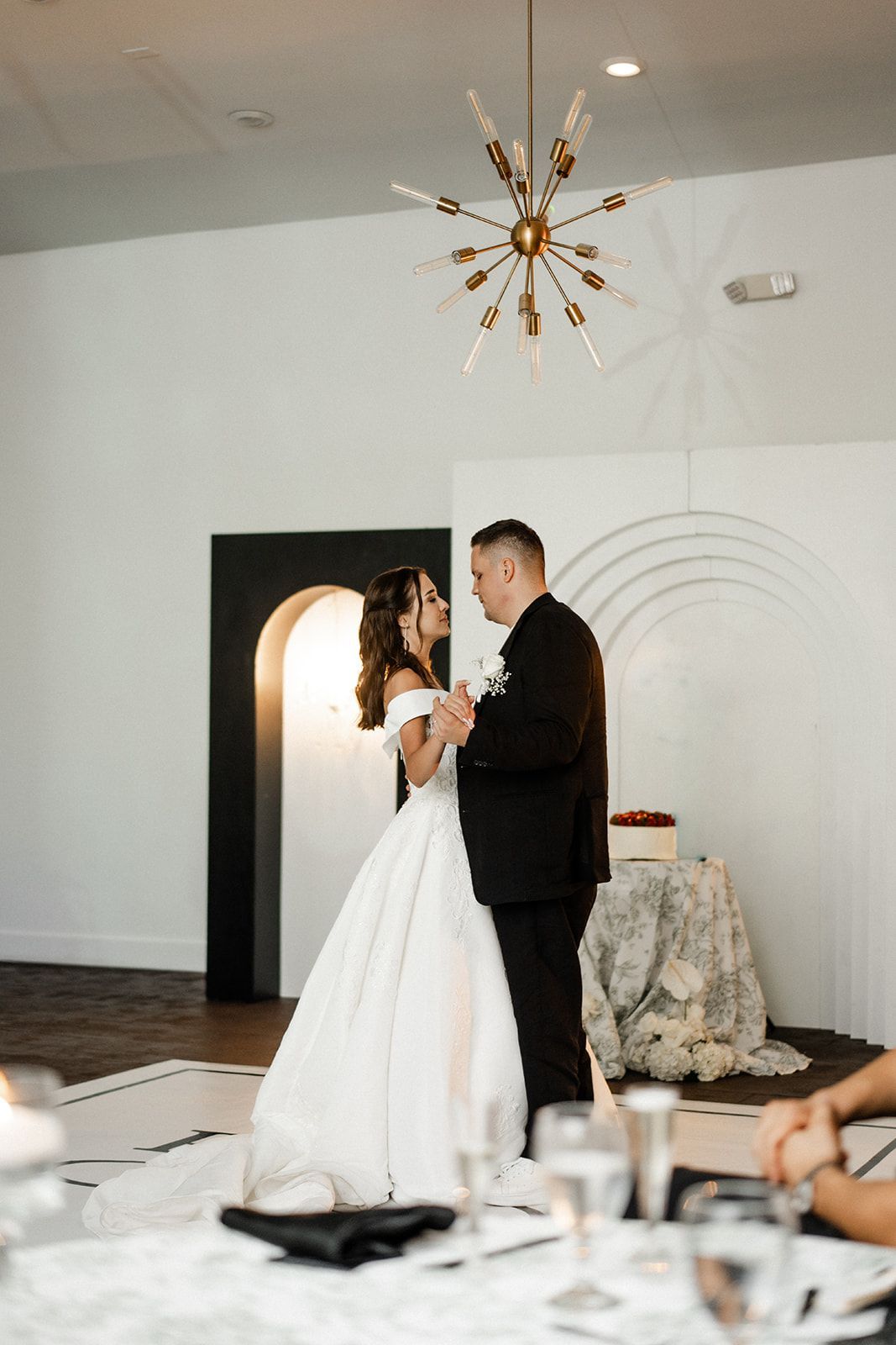 Bride and groom embrace at wedding reception, white dress, black suit, ornate chandelier, minimalist decor.
