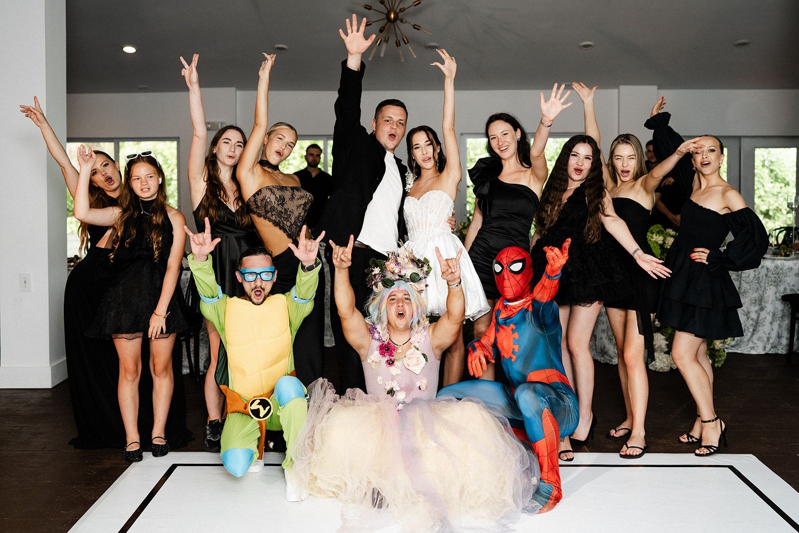 Group of people celebrating, arms raised. Various outfits, including costumes and formal wear, in a white-walled room with a dance floor.