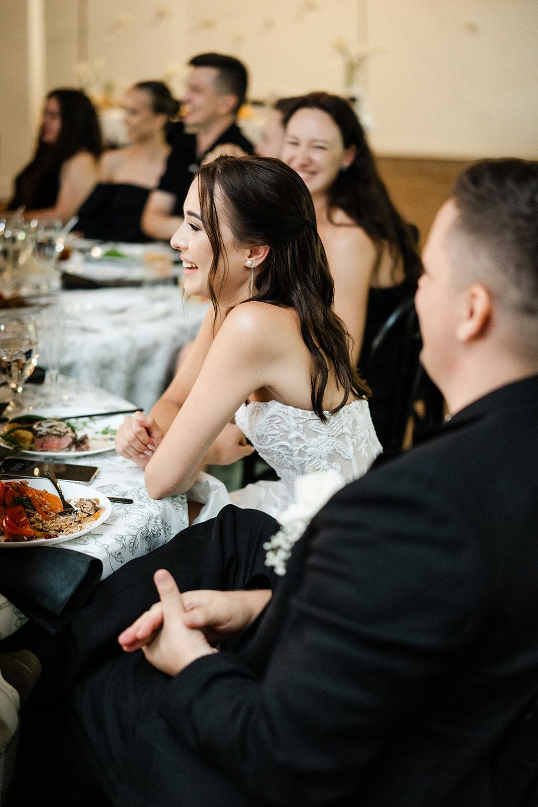 Bride smiles at a wedding reception, seated at a table. Others at the table look on.