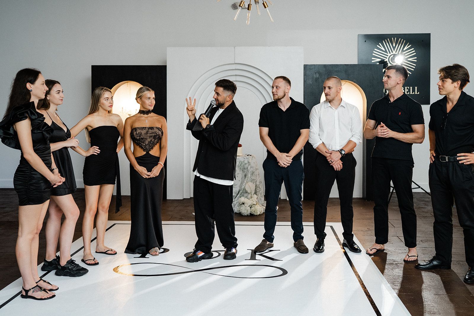 Group of people in black outfits gathered indoors. Man speaking, others listening on a white platform.