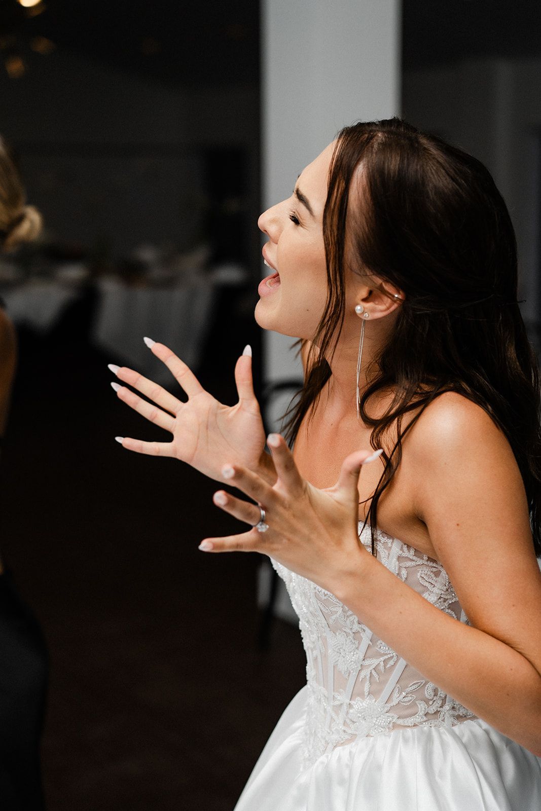 Woman in a white dress, singing with hands raised, mouth open wide, inside a venue.