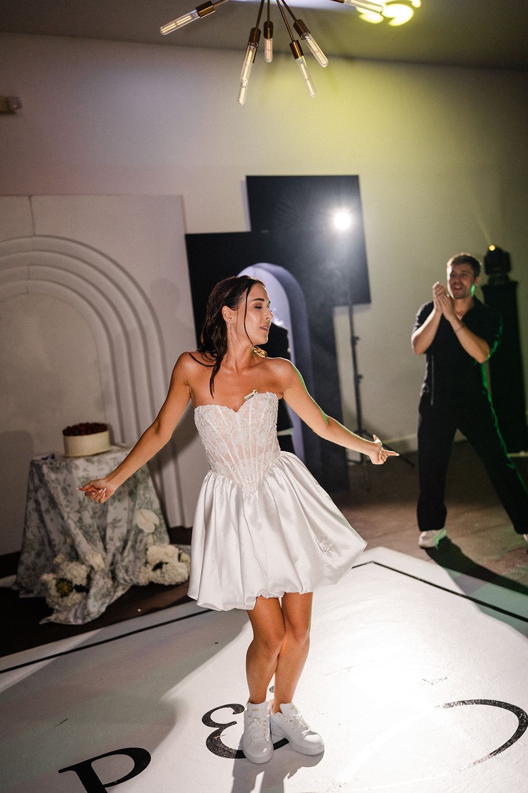 Woman in a white dress dances on a dance floor, another person claps. Elegant interior with cake, lights.