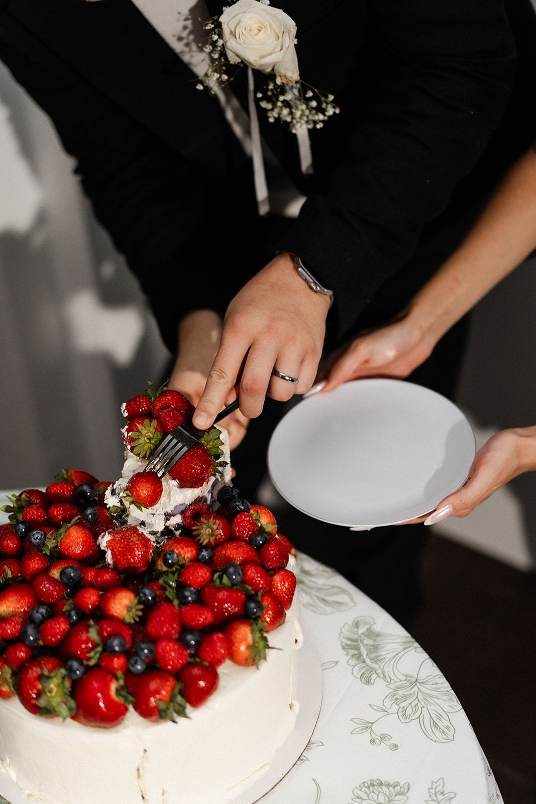 Couple cutting a heart-shaped cake, topped with strawberries and blueberries. A plate is held out to receive a slice.