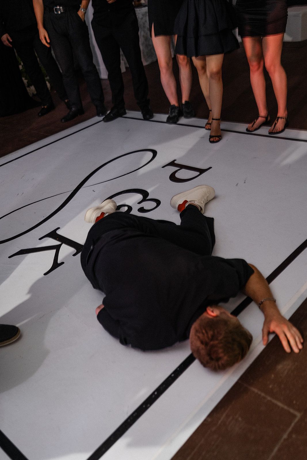 Person lies sprawled on a white dance floor with black border and logo. Other people stand around.