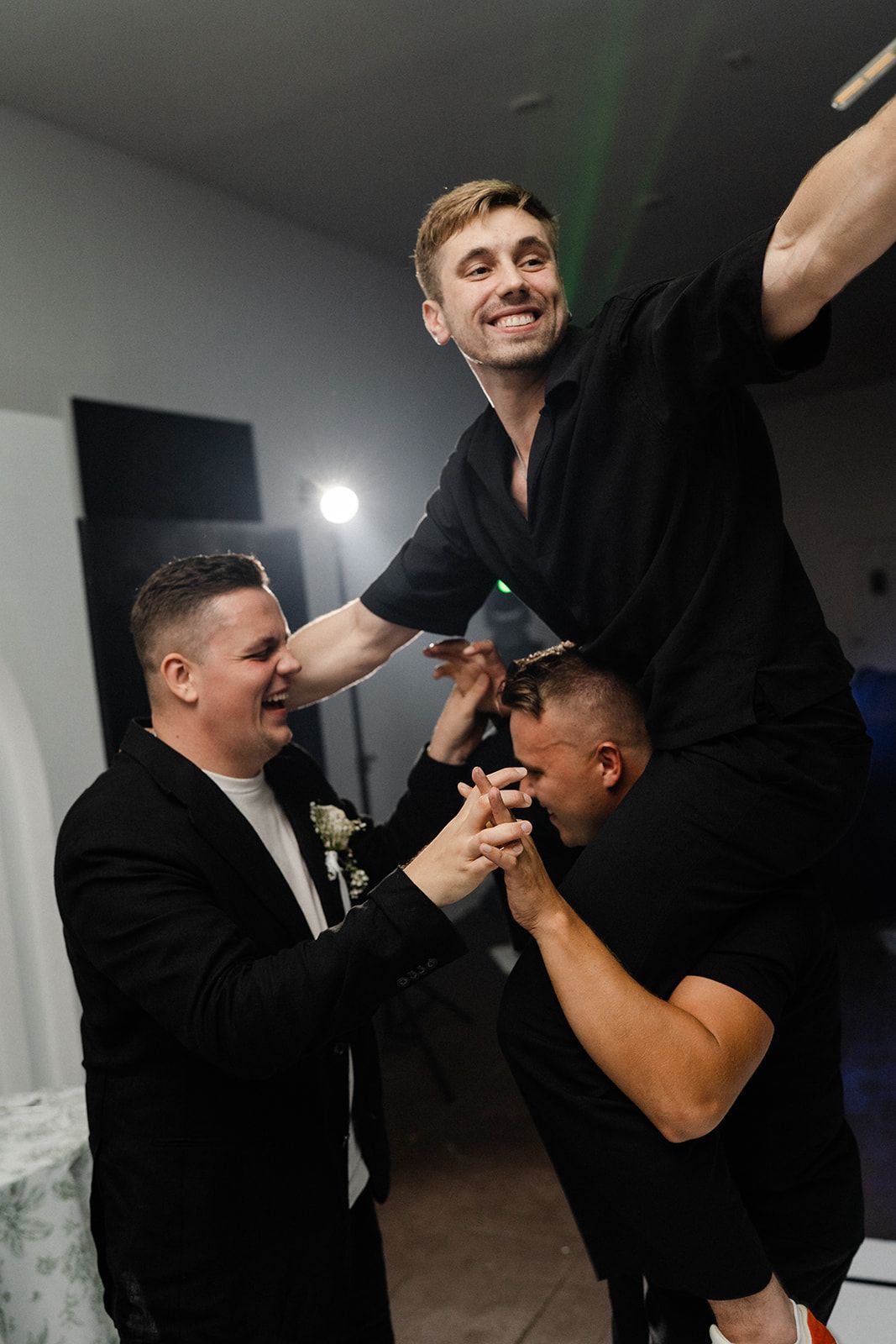 Three men celebrating. One man on another's shoulders, smiling. Another man supports them. Party setting.
