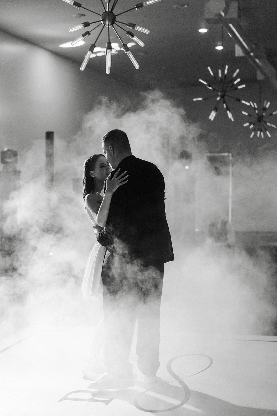 Couple dances in fog on a dance floor, beneath decorative lights. Black and white.