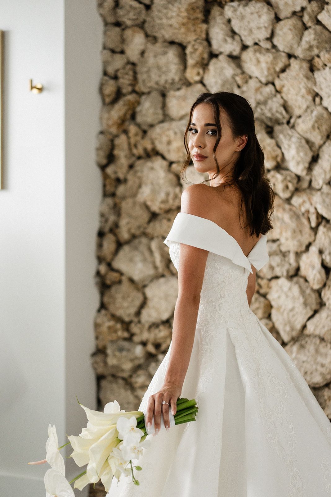 Woman in white off-shoulder wedding dress holding flowers, looking over her shoulder. Stone wall backdrop.