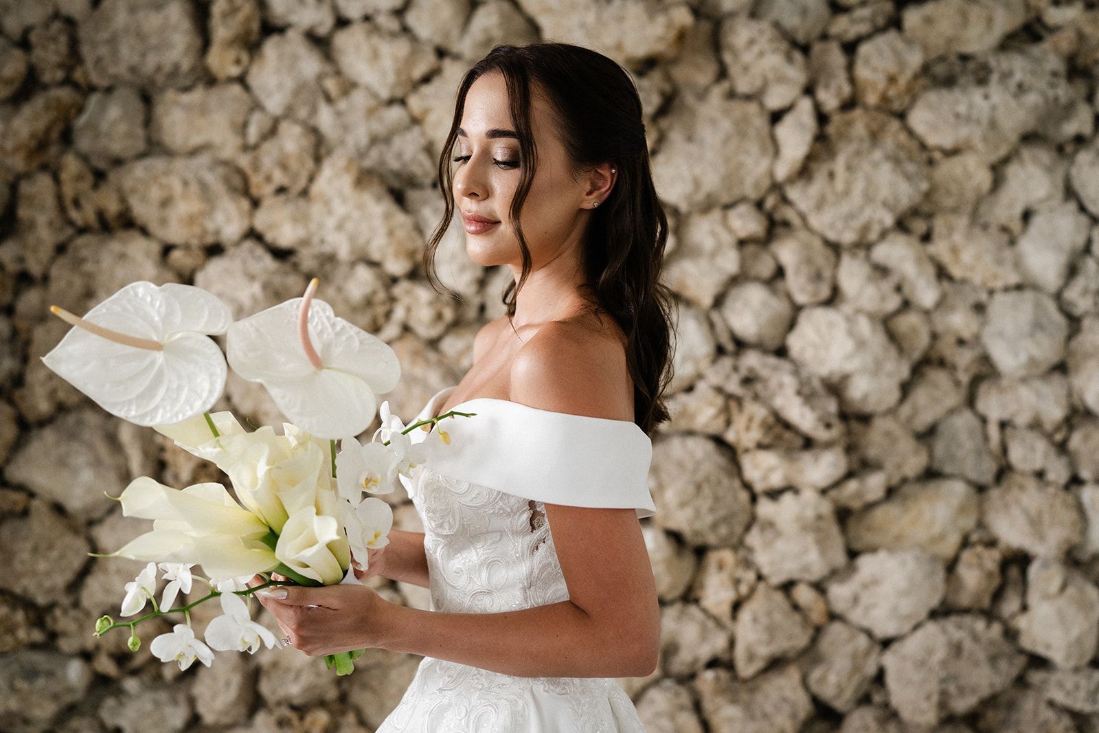 Woman in a white off-the-shoulder dress holding a bouquet of white flowers in front of a stone wall.