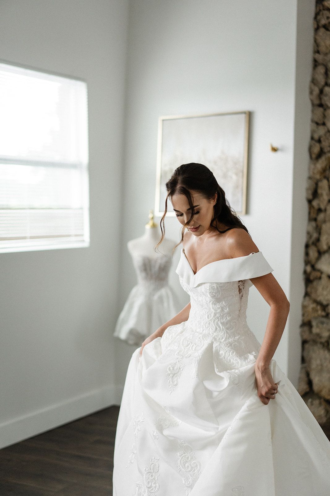 Woman in a white wedding dress adjusting the skirt, standing in a room with a dress form.