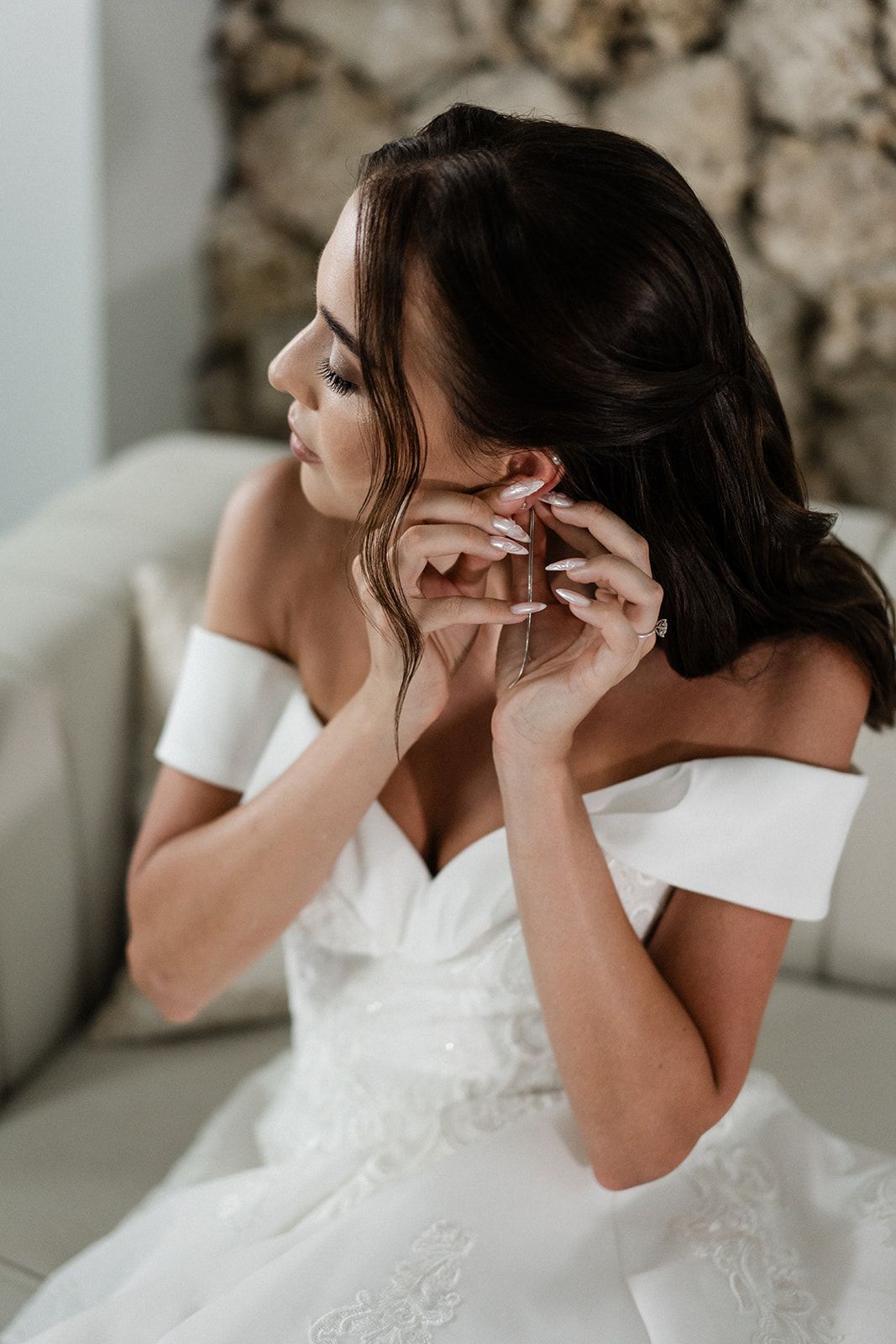 Bride in white off-the-shoulder dress, putting on earrings, indoors.