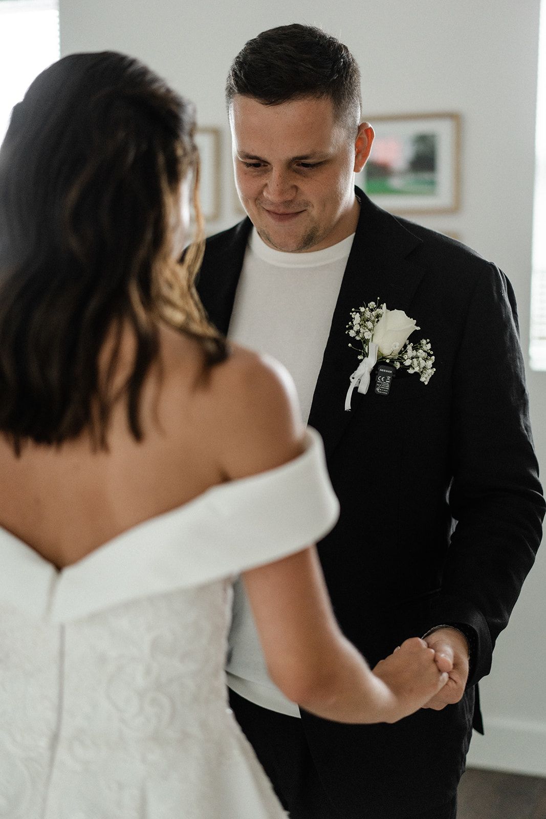 Bride and groom holding hands, smiling. The groom is wearing a black suit and has a boutonniere. The bride is wearing a white dress.