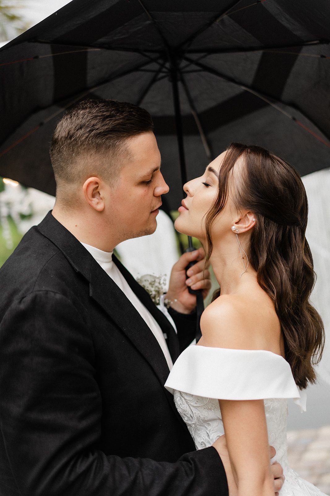 Couple under an umbrella, bride in white dress, groom in black suit, about to kiss.