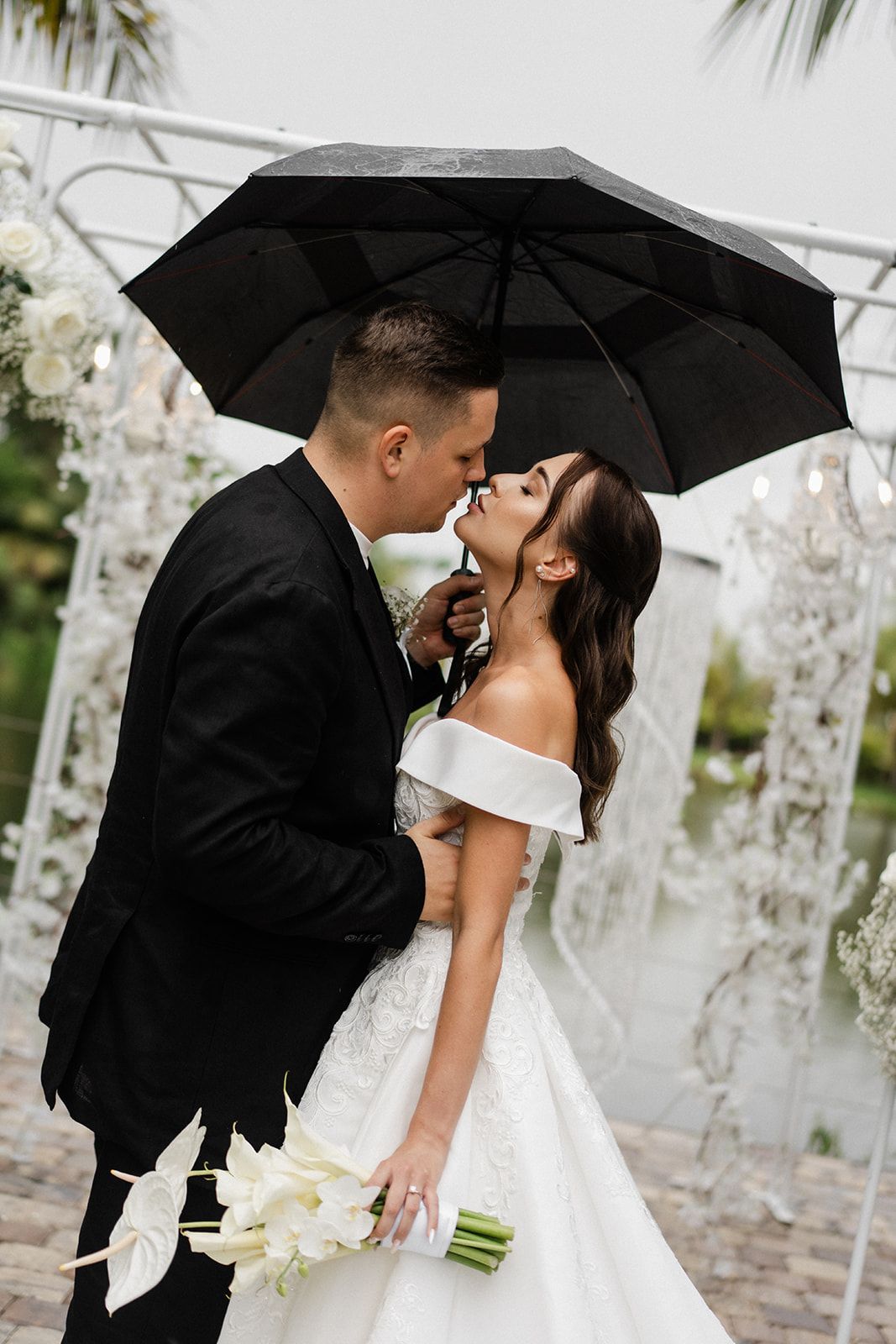 Bride and groom kissing under a black umbrella. She wears a white gown, he a black suit. Rain is falling.