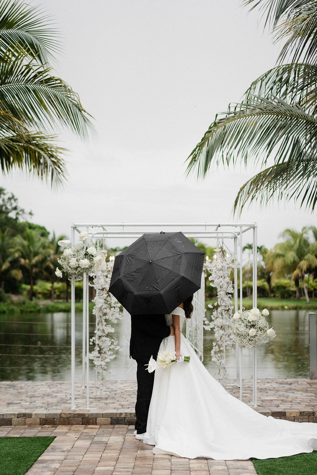 Bride and groom under a black umbrella, kissing near a pond, floral archway.