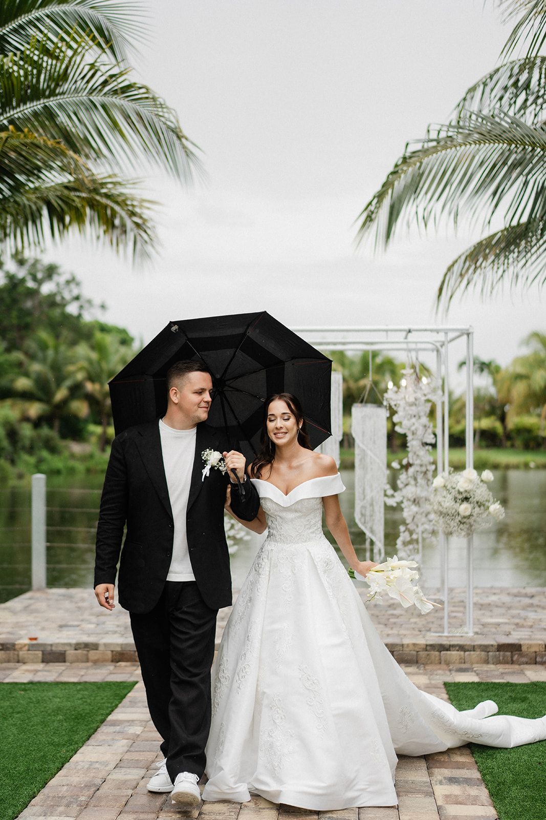 Couple walking under a black umbrella on a brick path, with a wedding backdrop and pond in the background.