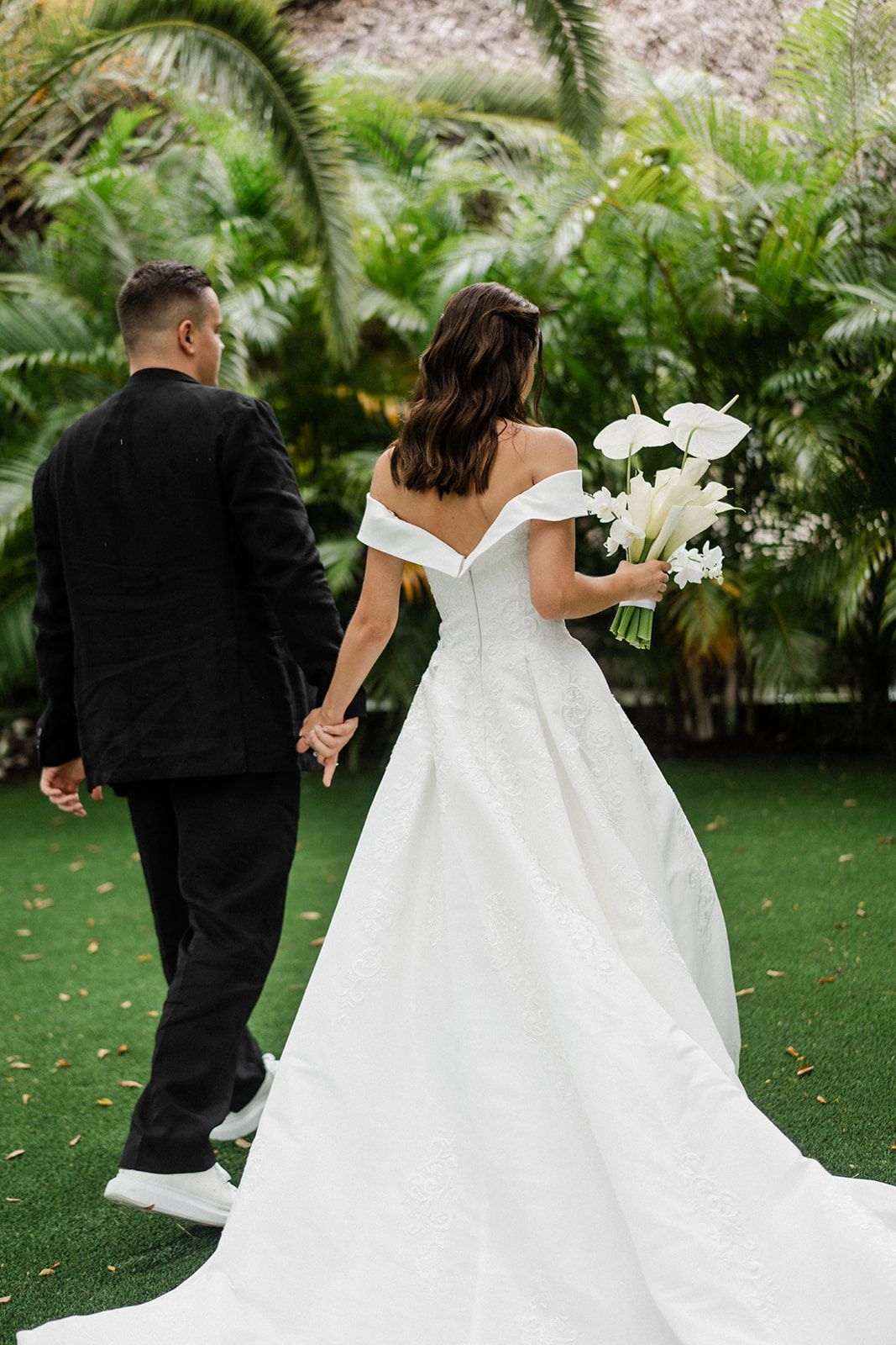 Bride and groom holding hands, walking away. Bride in white dress, groom in black suit. Outdoors with greenery.