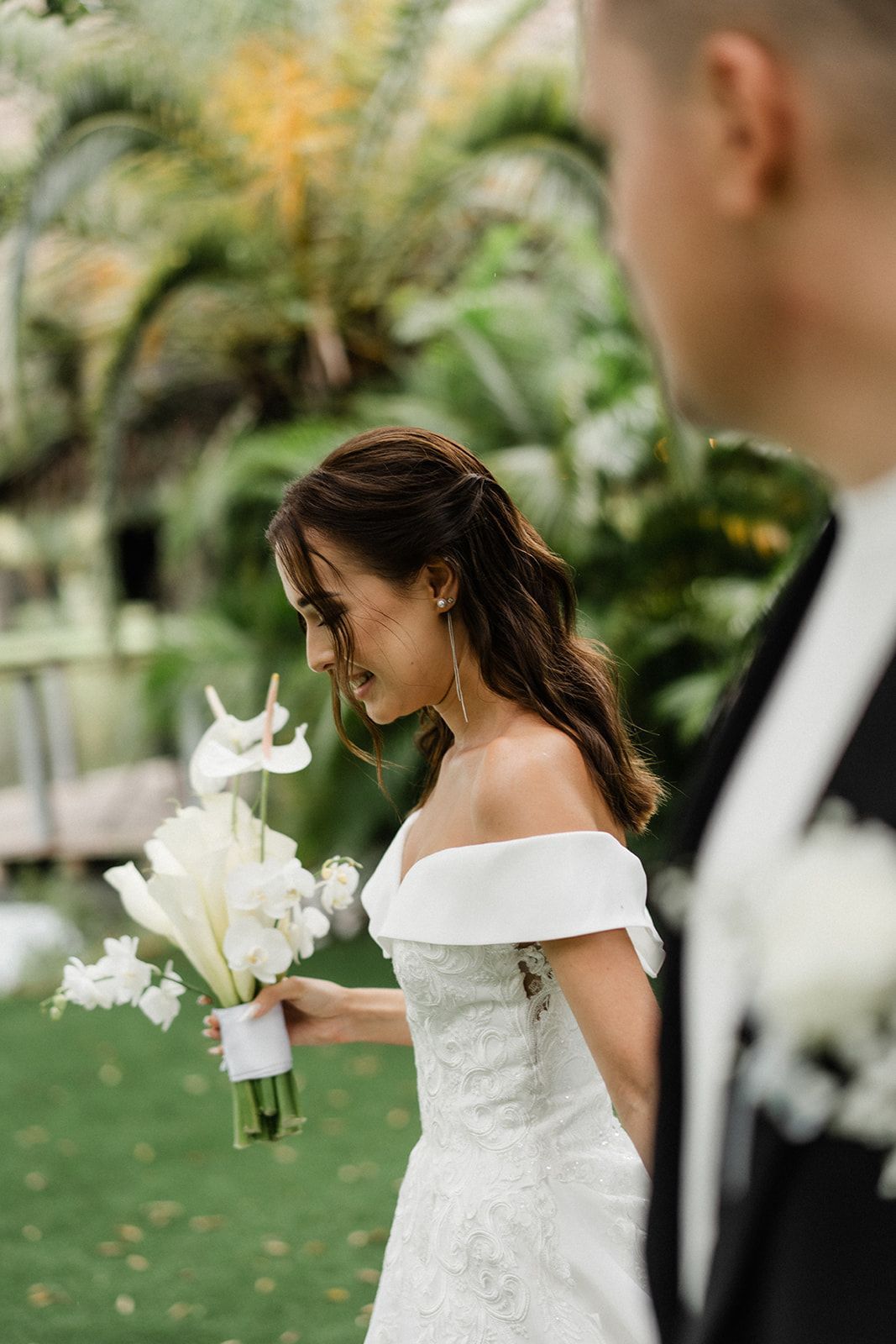 Bride in a white off-shoulder dress holding a bouquet, looking down. Groom in a black suit partially visible. Outdoor wedding.