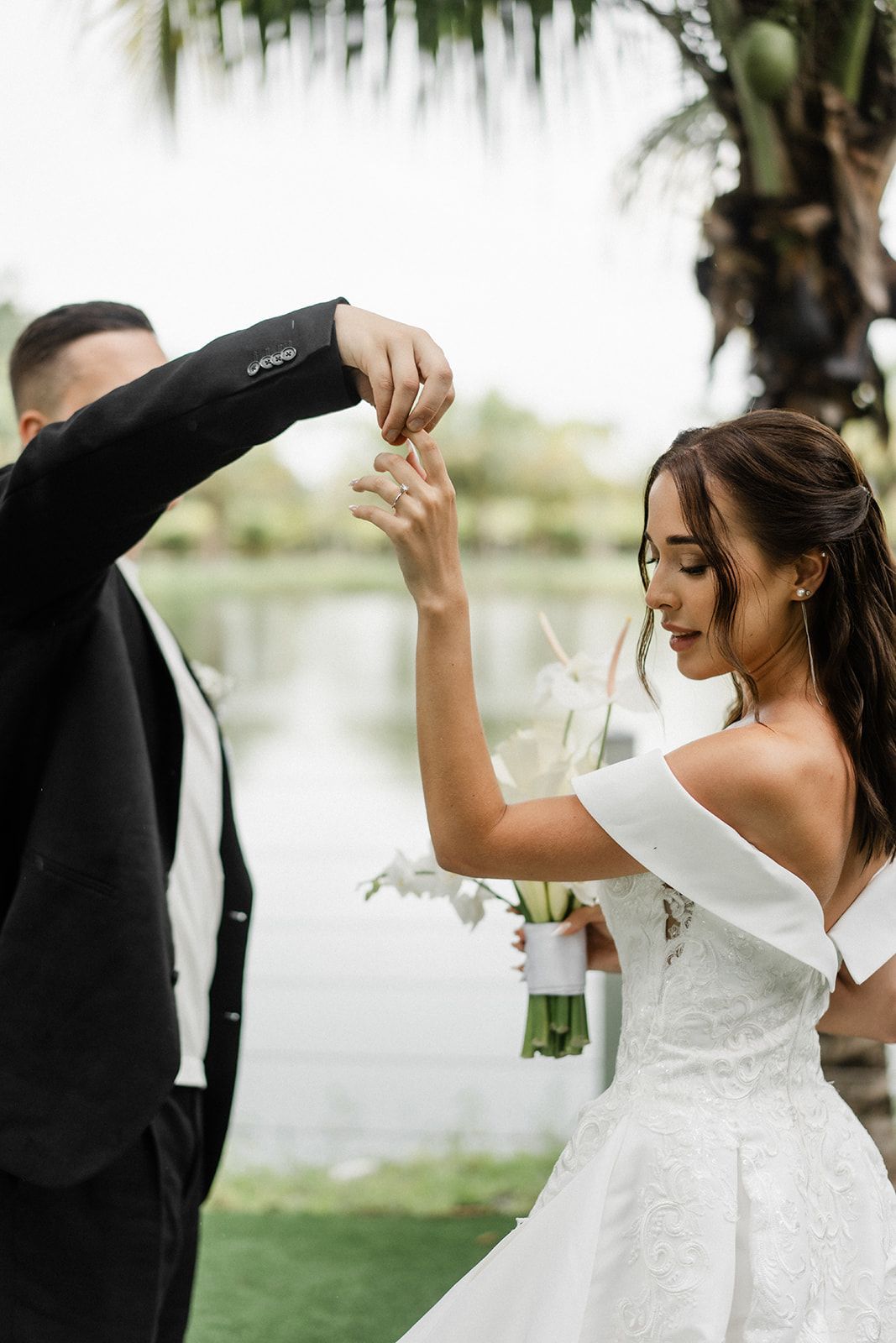 Groom placing ring on bride's finger, outdoors. Bride in white dress, holding bouquet. Lake in background.