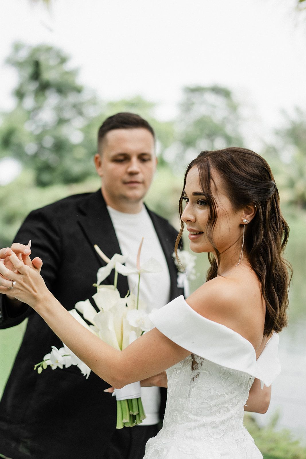 Bride and groom dancing outdoors; bride in white dress holding flowers, groom in black suit.