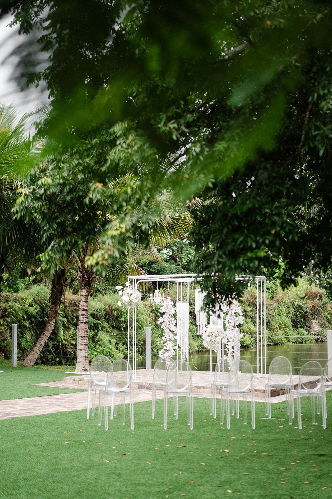 Ceremony setup with clear chairs on green lawn, white arch, and trees near water.