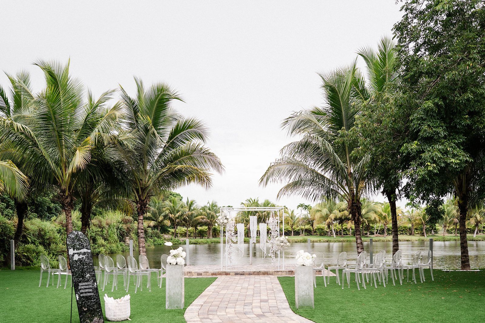 Wedding ceremony setting with water feature, pathway, palm trees, and chairs on green grass.