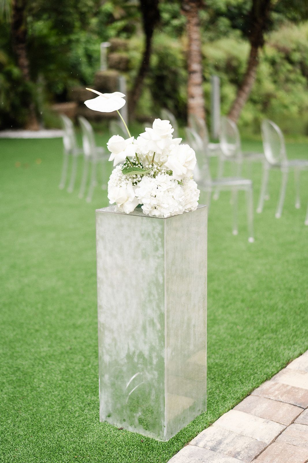 White flowers on a clear pedestal in a grassy outdoor setting with chairs.