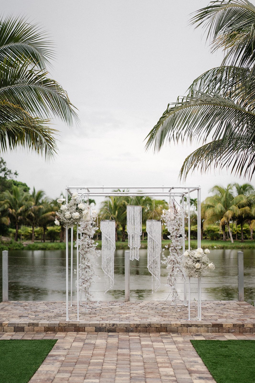 White wedding arch decorated with flowers and hanging fabric against a lake and palm trees.