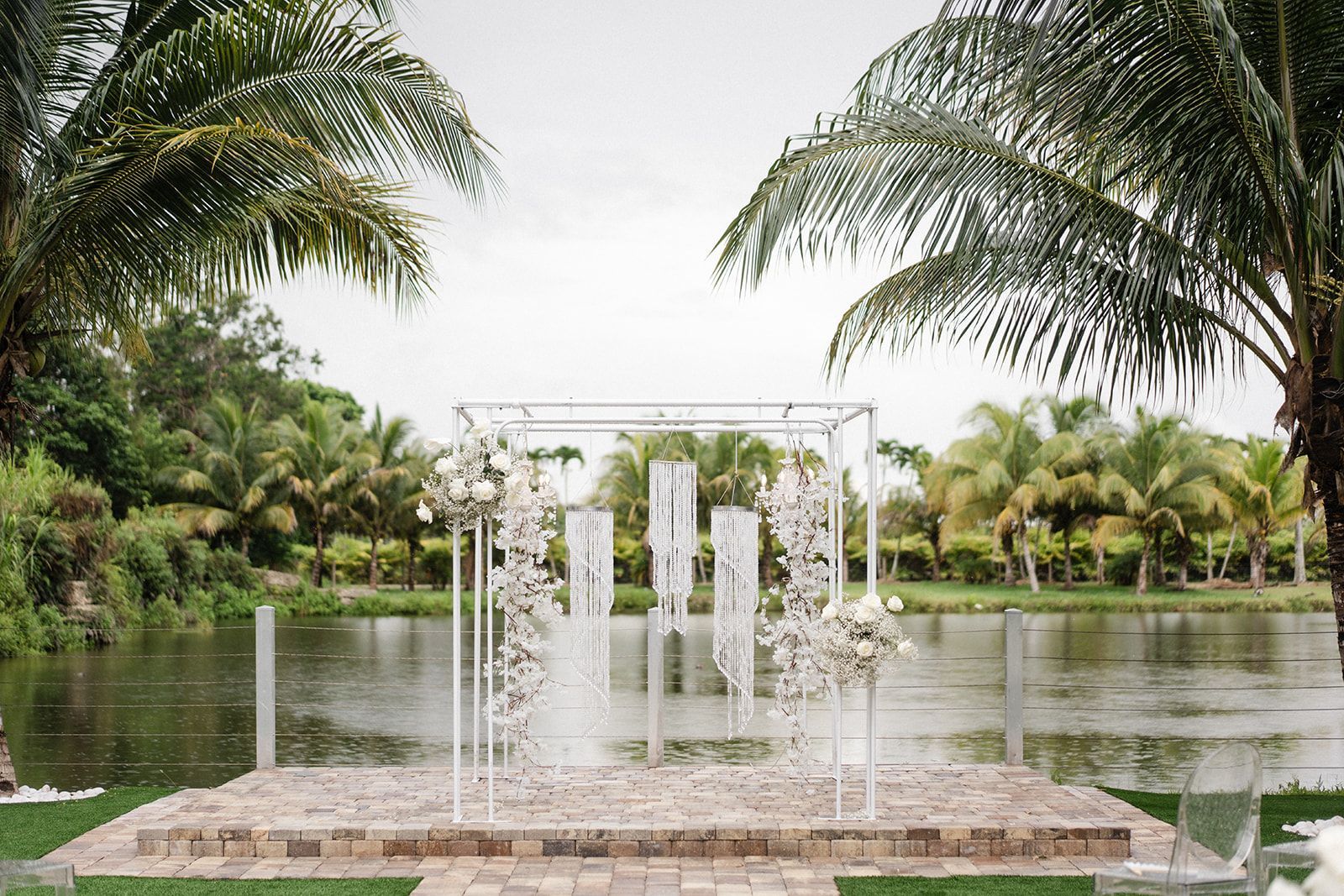 Wedding ceremony setup with white floral arch over brick platform, by a lake with palm trees.
