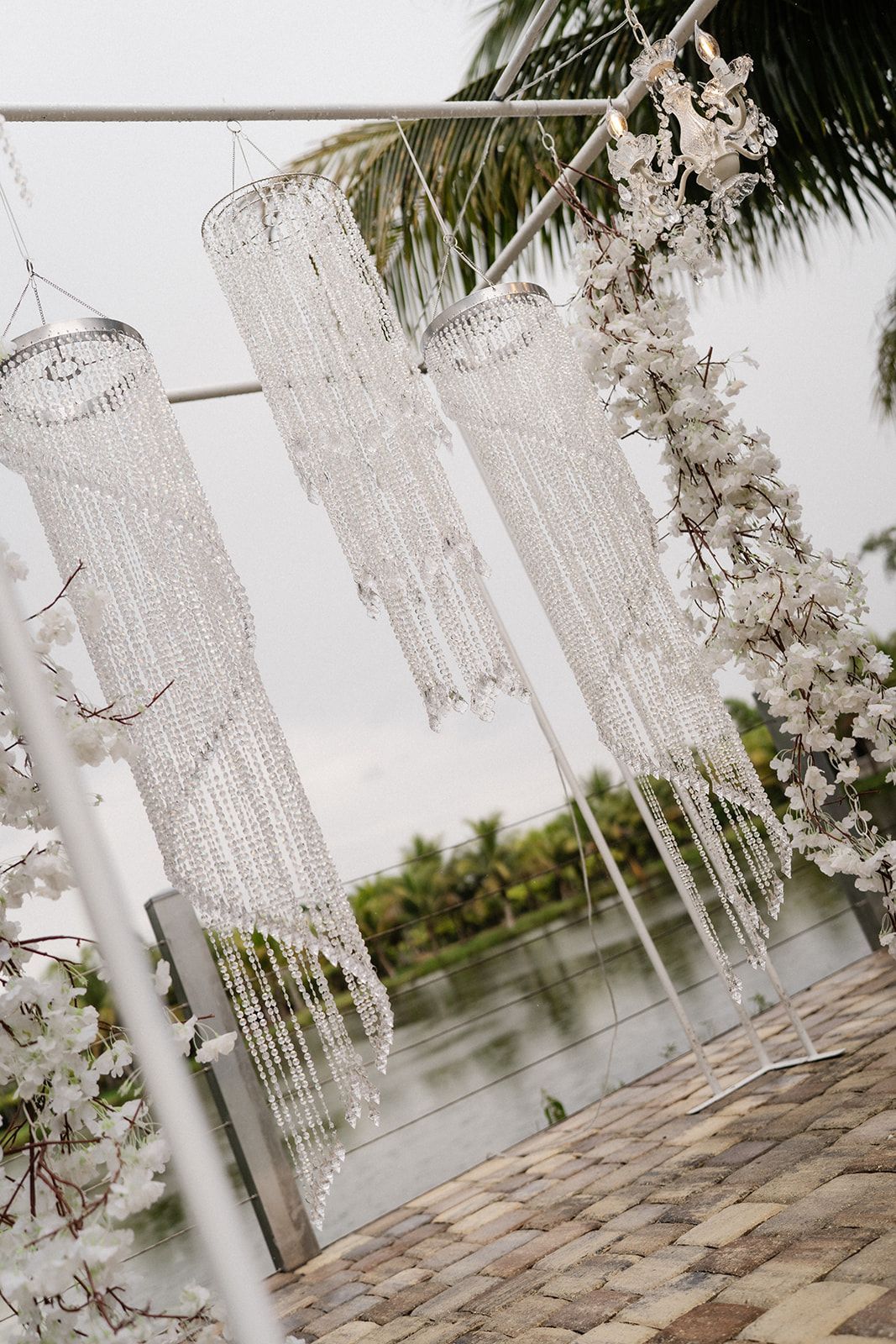Three beaded chandeliers hang from a metal arch, with white flowers. Water and trees in the background.