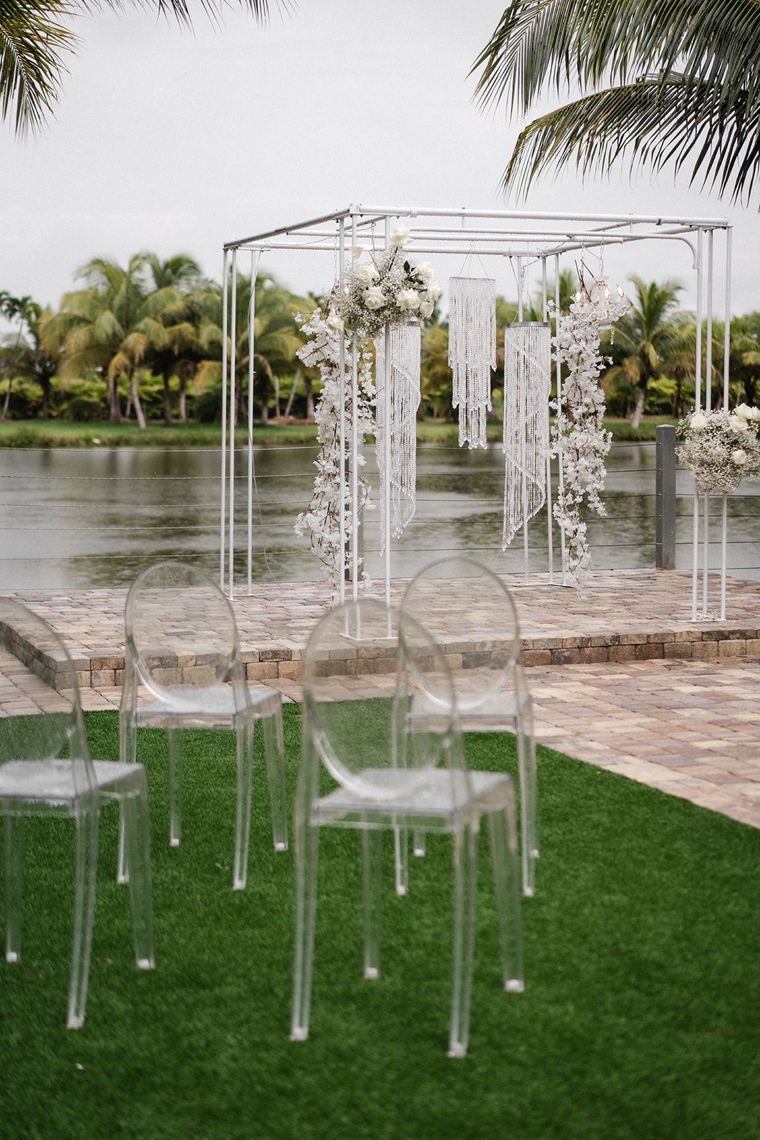 Wedding ceremony setup with clear chairs on green grass by a lake, white floral decorations.