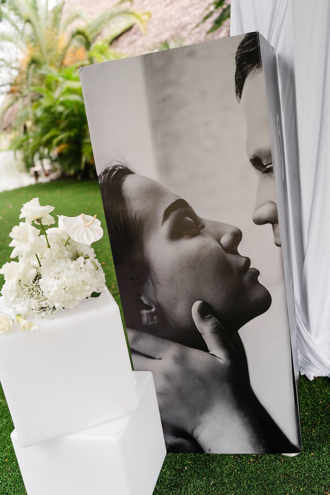 Black and white photo of a couple on a rectangular display. The woman's face is angled up, man touches her face. White flowers on stacked blocks.