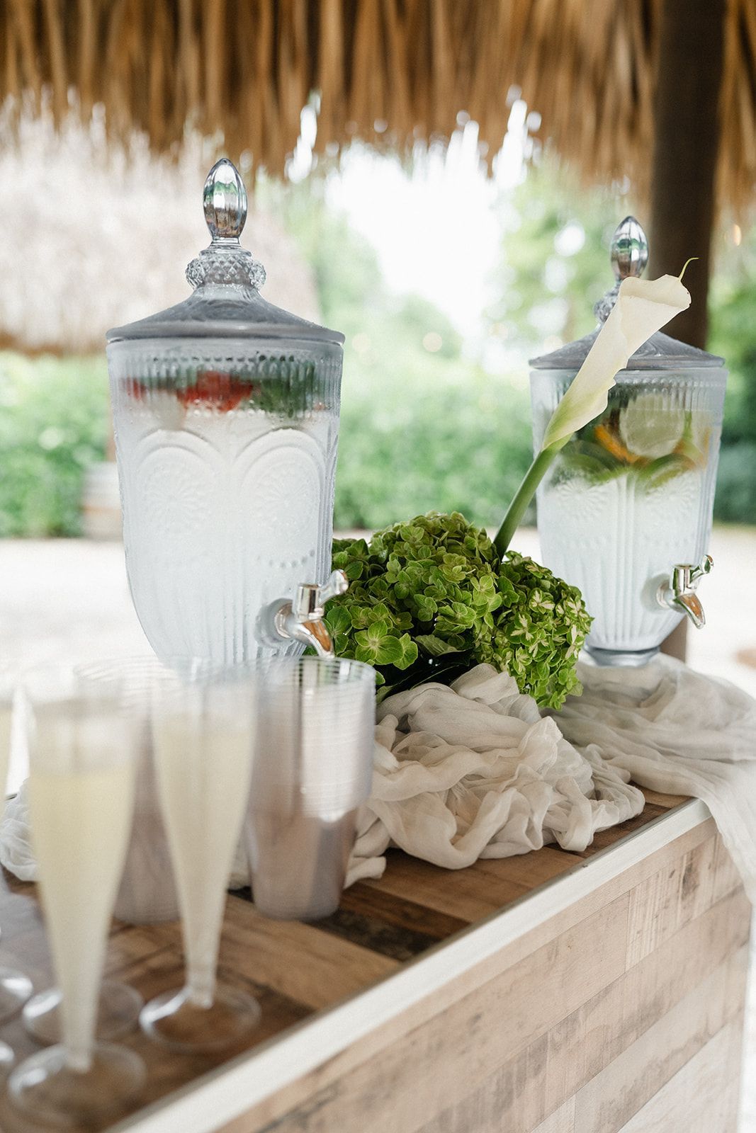 Beverage station with two glass drink dispensers, greenery, and champagne flutes.