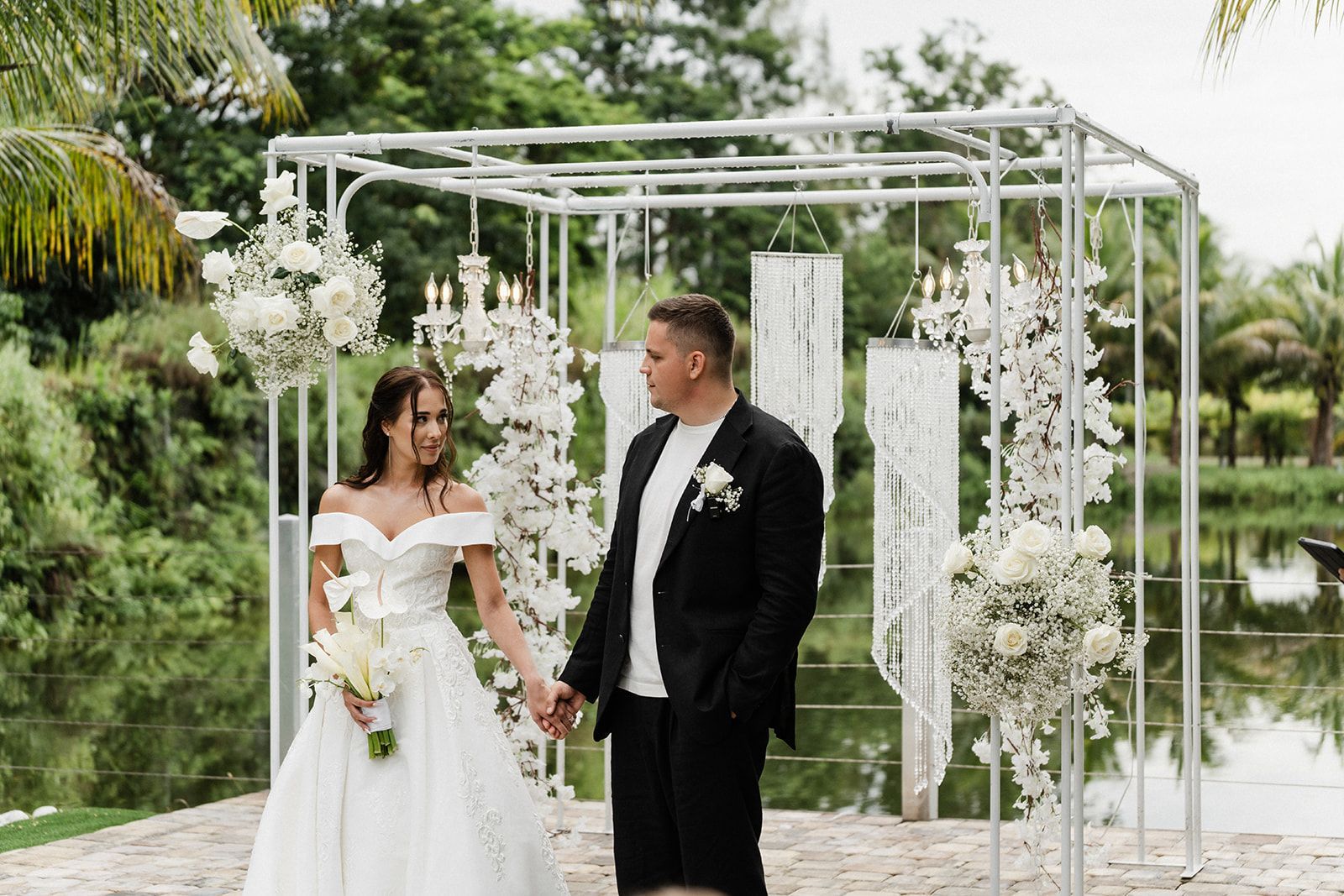 Bride and groom holding hands, standing near decorated wedding arbor by water.
