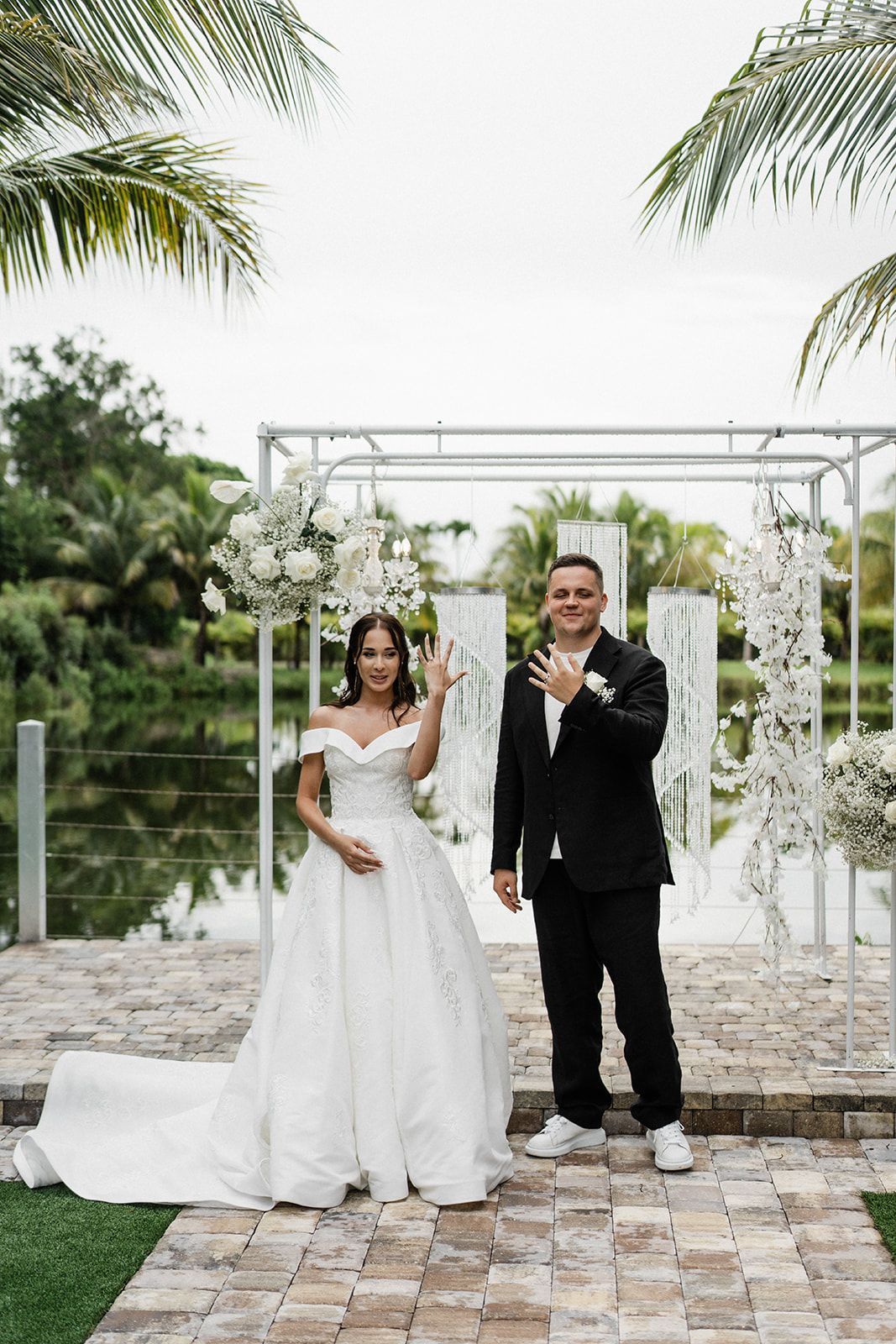 Bride and groom posing by a wedding arbor; bride in white gown, groom in suit and sneakers.
