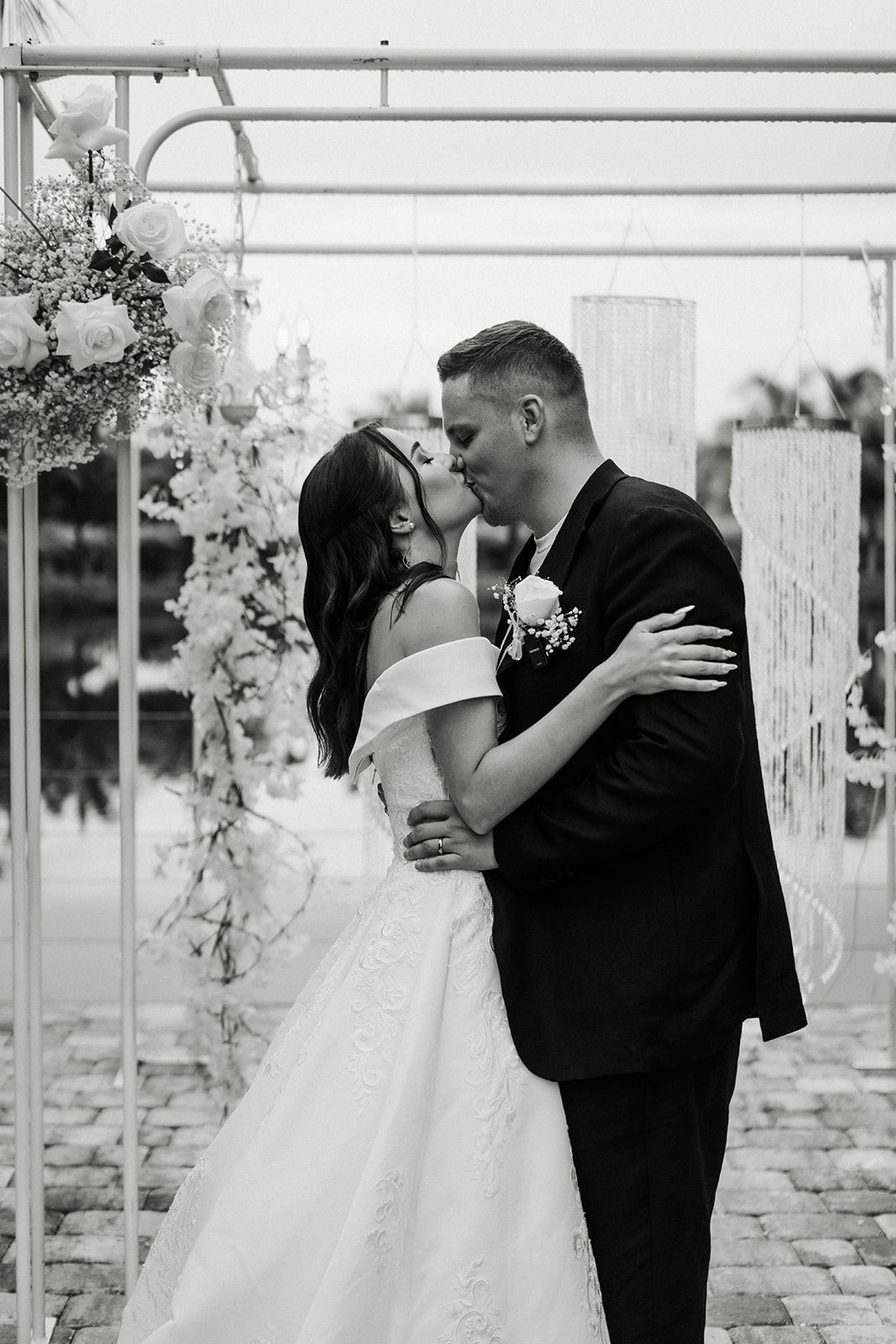 Newlyweds kissing under a white archway decorated with flowers. The bride wears a white gown, and the groom wears a black suit.