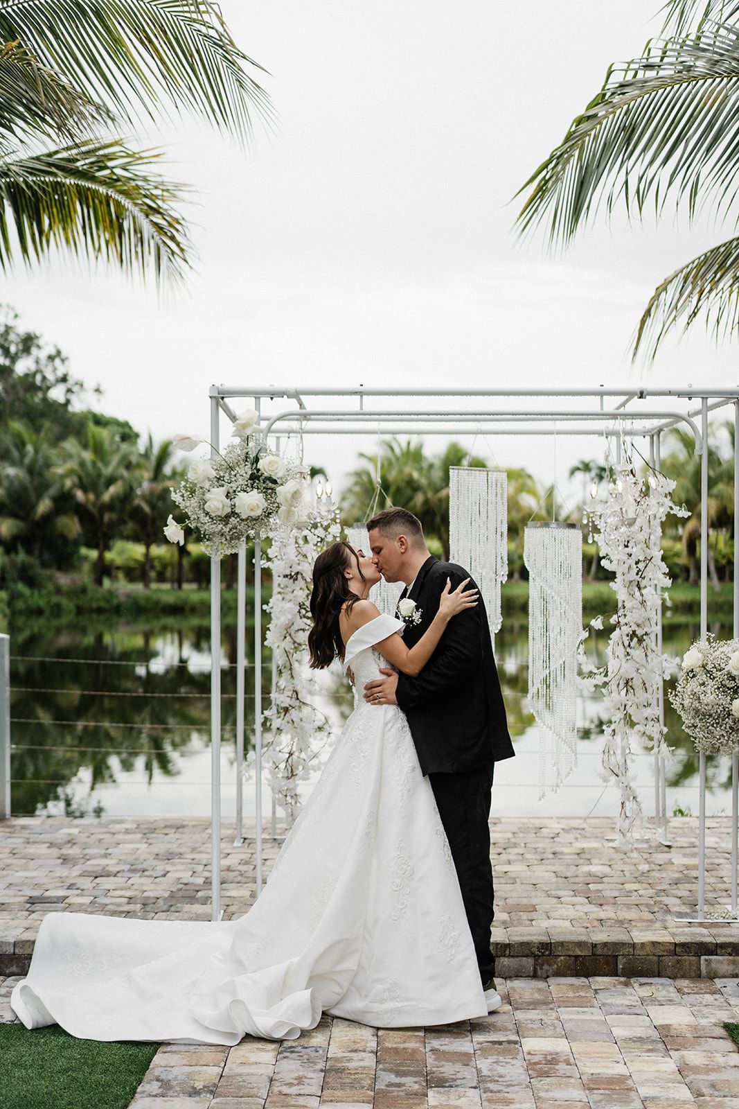 Bride and groom kiss at outdoor wedding ceremony by a reflective lake.