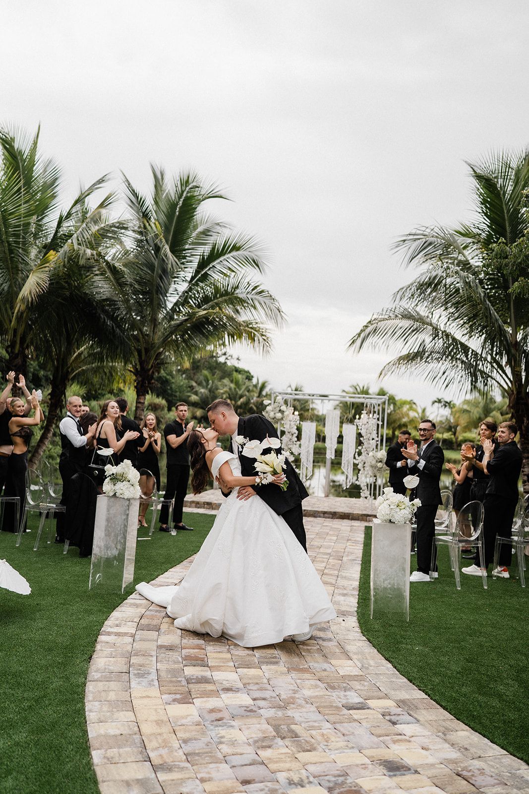 Newlyweds kiss on brick path, cheered by wedding guests outdoors.